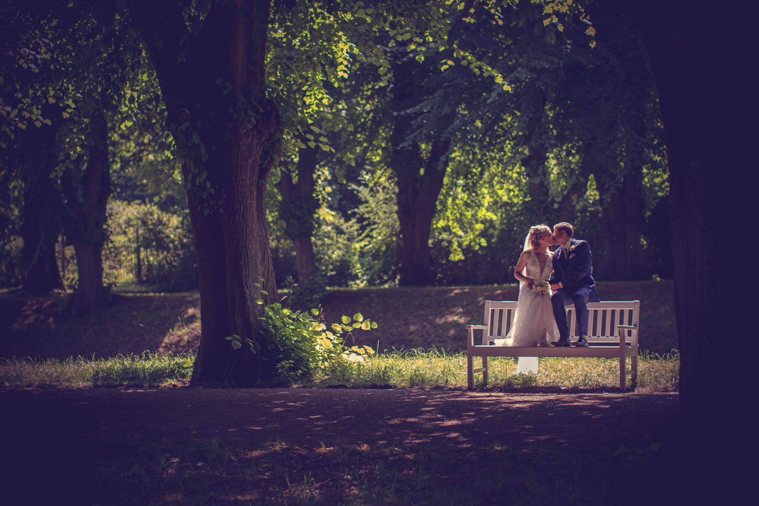 Romantische Aufnahme eines Brautpaares, das auf einer weißen Parkbank in der Allee im Park von Schloss Bothmer sitzt. Das malerische Sommerlicht schafft eine verträumte Atmosphäre. Romantische Aufnahme eines Brautpaares, das auf einer weißen Parkbank in der Allee im Park von Schloss Bothmer sitzt. Das malerische Sommerlicht schafft eine verträumte Atmosphäre.