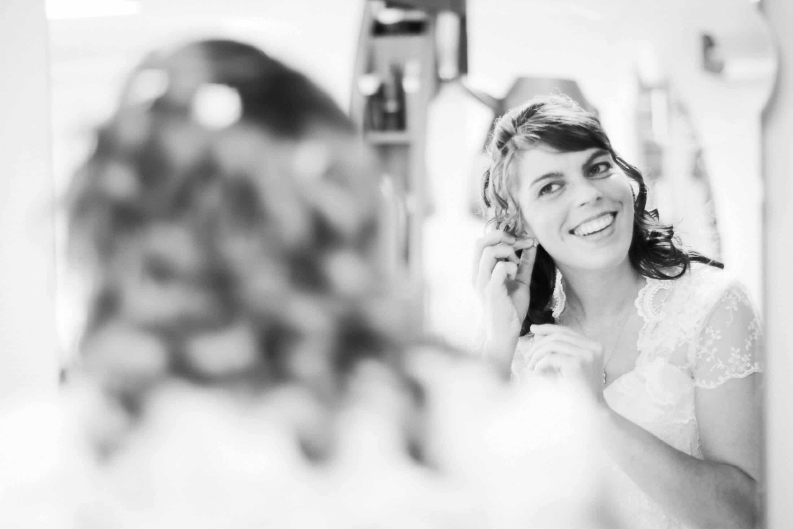 Braut steckt sich Ohrringe an, Spiegelaufnahme im Hochzeitskleid – elegantes Schwarz-Weiß-Foto vom Getting Ready