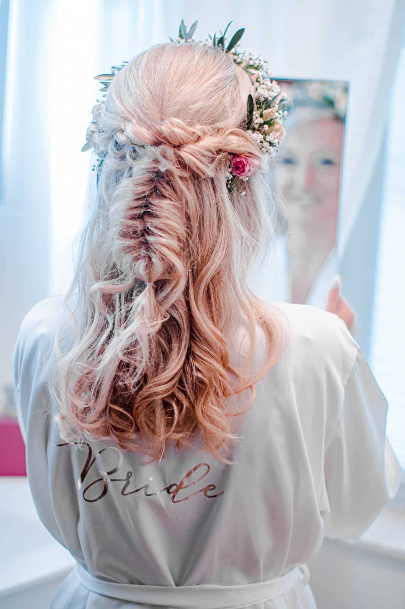 Braut von hinten in Morgenmantel mit Blumen im Haar, aufgestylte Hochzeitsfrisur vor dem Spiegel – detailverliebtes Getting Ready-Foto