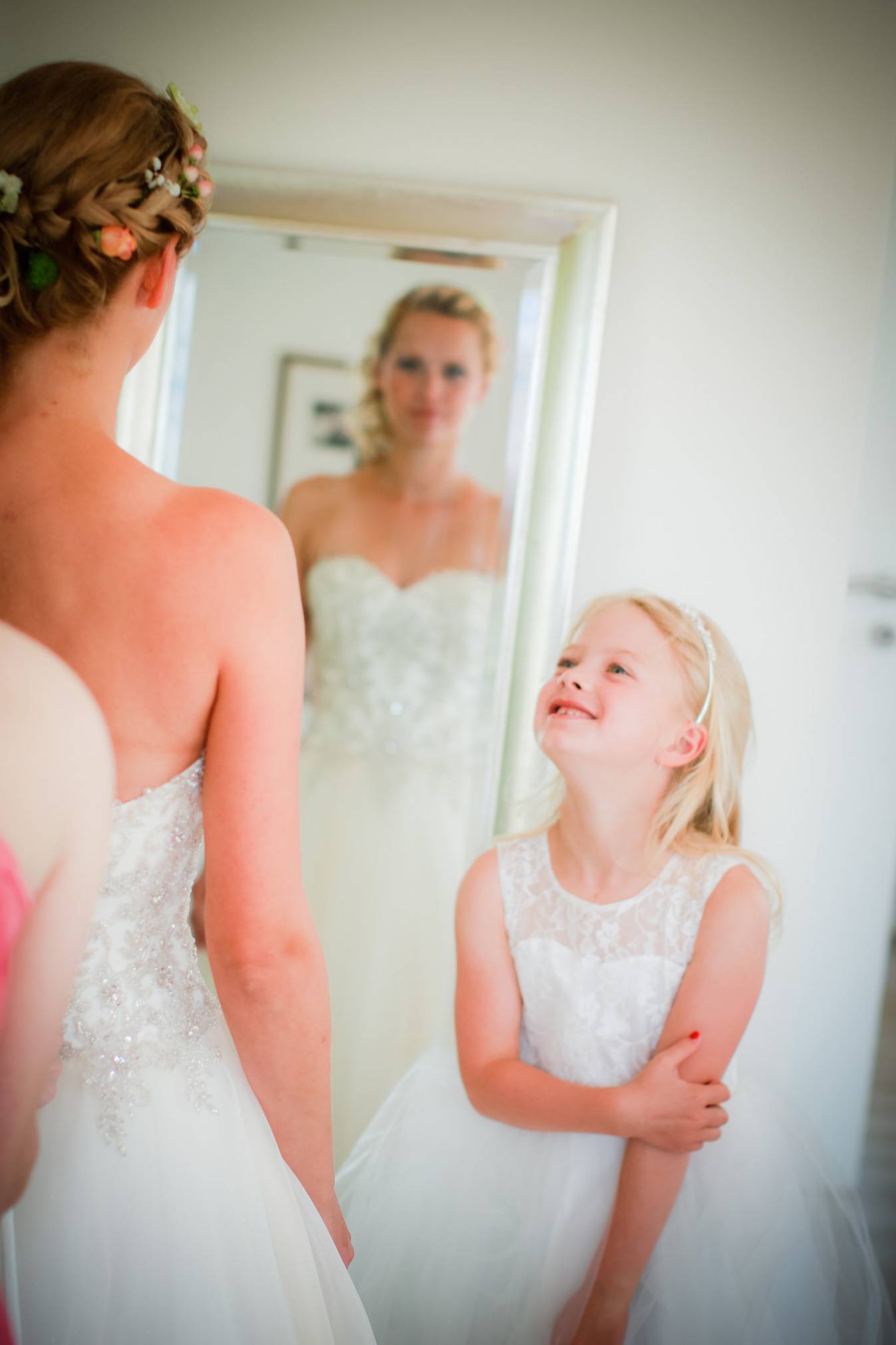 Braut mit Blumen im Haar sieht sich im Spiegel, Blumenmädchen im weißen Kleid bewundert sie – intimer Hochzeitsmoment am Morgen