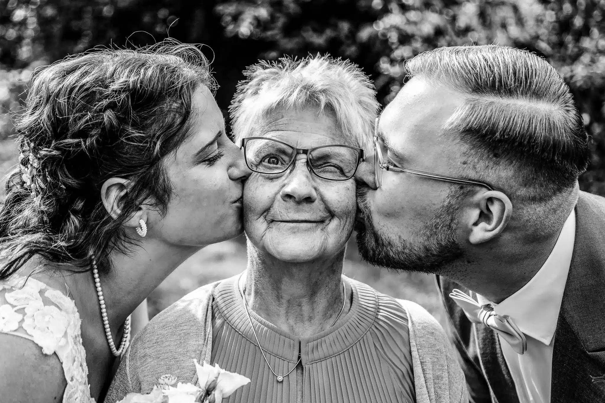 Ein herzliches Generationenfoto in Schwarz-Weiß: Braut und Bräutigam geben ihrer glücklich zwinkernden Großmutter gleichzeitig einen Kuss auf die Wange. Ein Bild voller Liebe und Humor.