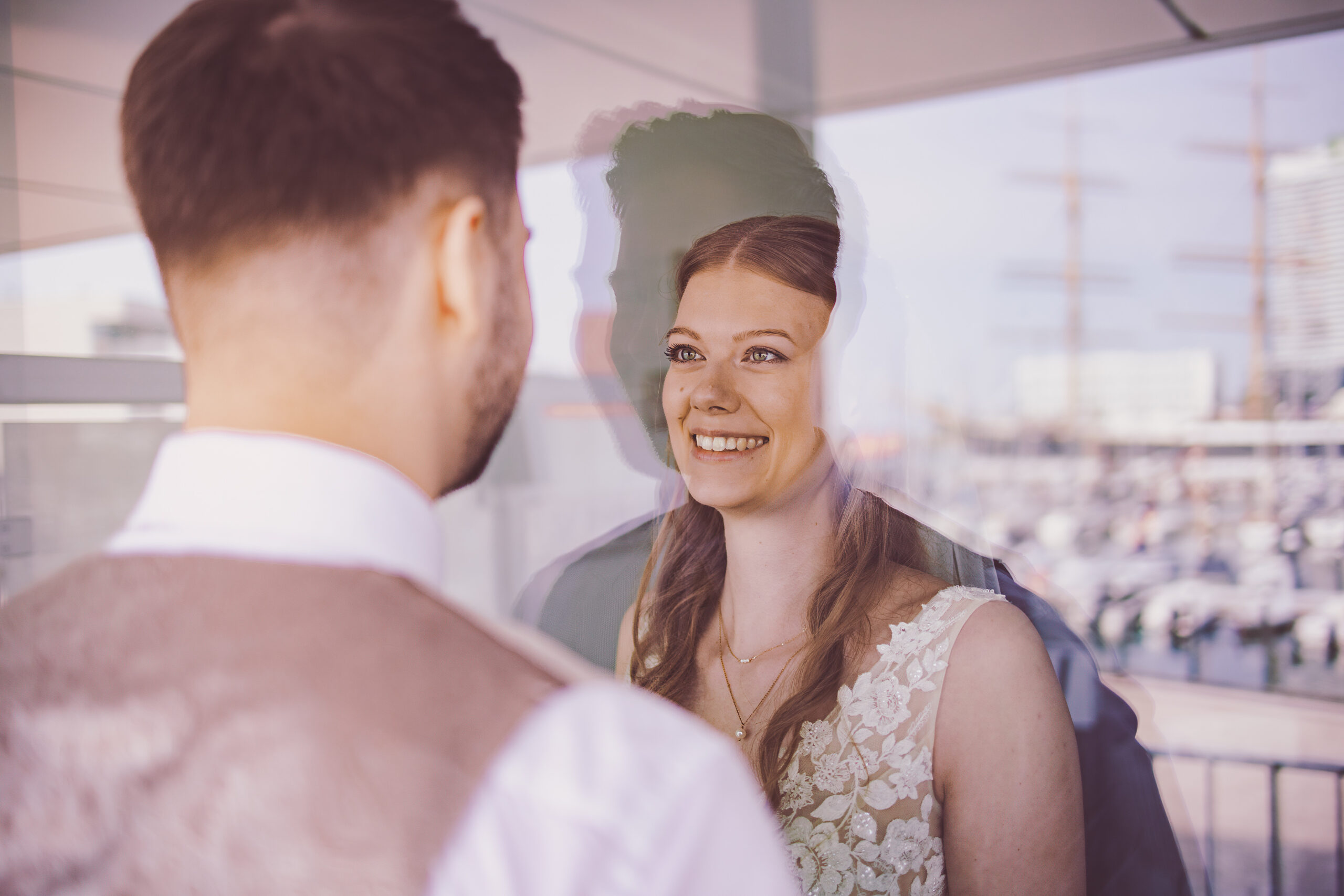 _MG_7289-Bearbeitet Braut und Bräutigam blicken sich mit einem Lächeln durch eine Fensterscheibe auf dem Priwall an – romantischer First Look mit maritimer Kulisse im Hafen von Travemünde