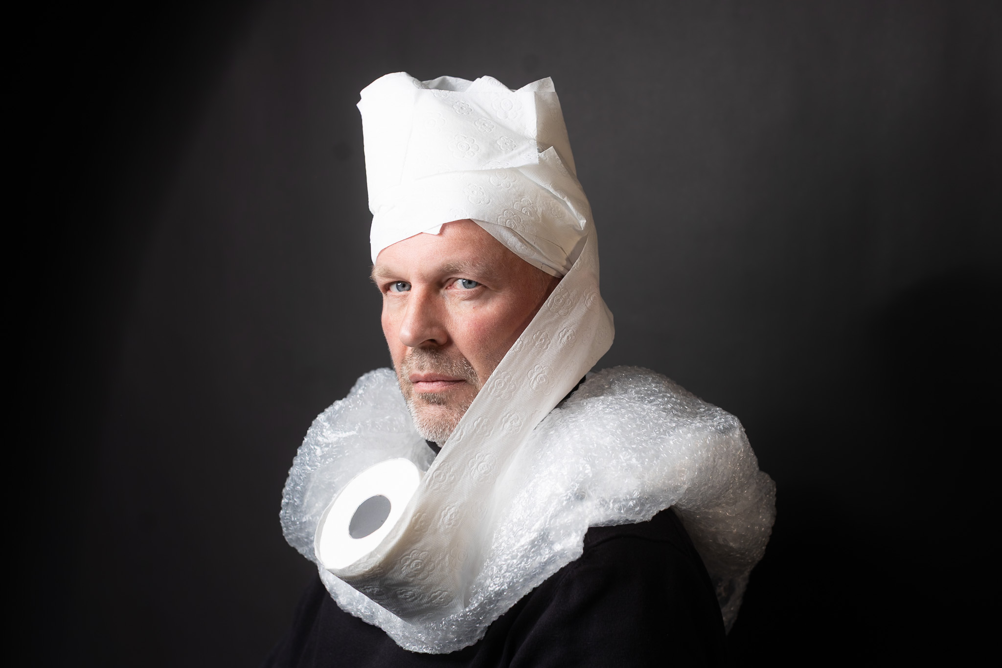 Tutorial: Verwandle dein Selfie in ein Rembrandt-Meisterwerk 1 Unbearbeitetes Porträtfoto eines Mannes mit selbstgemachtem Hut aus Toilettenpapier und Kragen aus Luftpolsterfolie vor dunklem Hintergrund – Ausgangsbild für Rembrandt-Look.