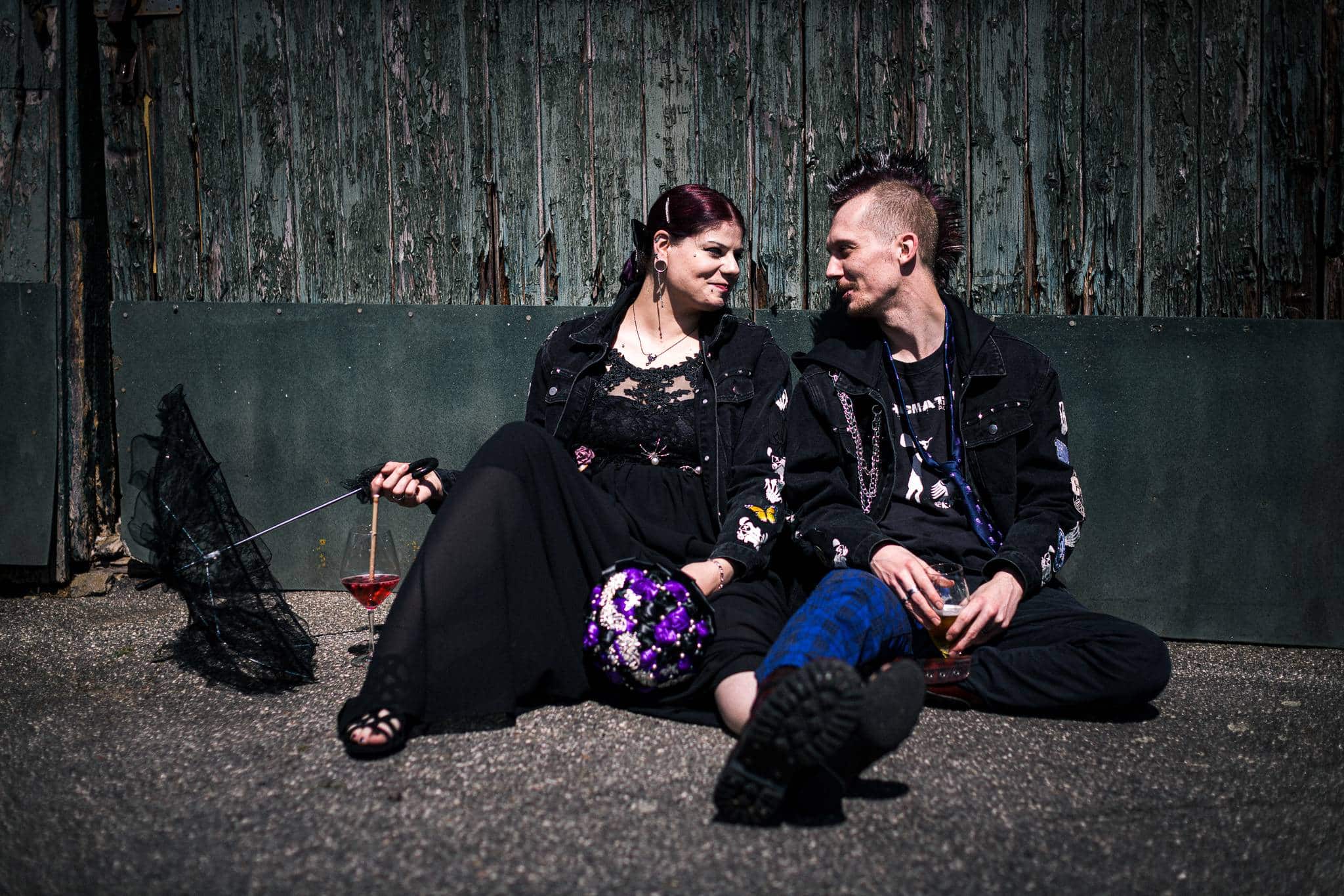 Ein alternatives Brautpaar im Punk-Stil sitzt entspannt auf dem Boden vor einer alten Holzwand. Sie schauen sich lächelnd an und halten Getränke in den Händen. Neben der Braut liegen ein schwarzer Spitzenschirm und ihr lila-weißer Brautstrauß. Die Aufnahme fängt einen ruhigen, authentischen Moment der Zweisamkeit ein.