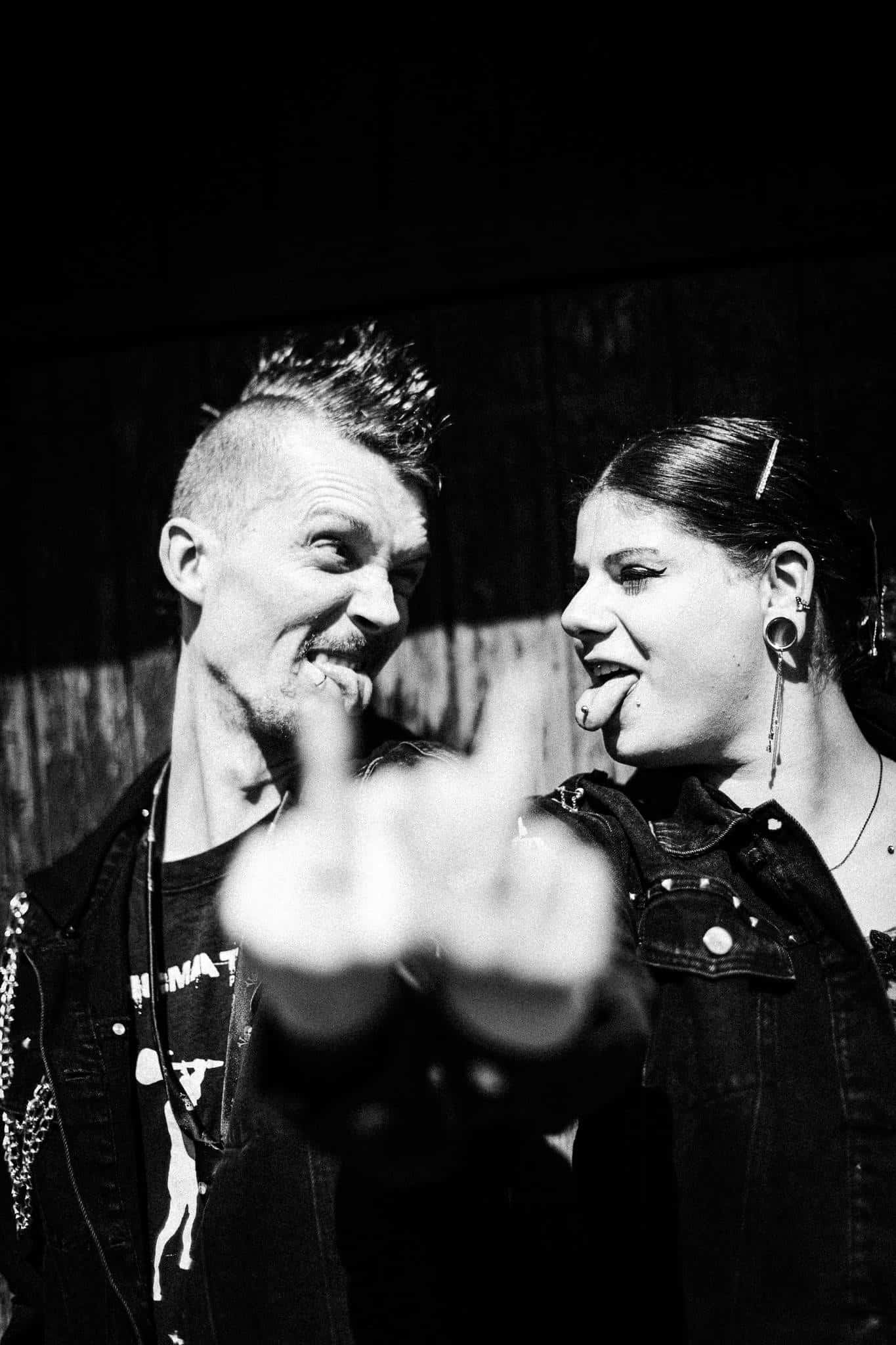 Ein provokantes Schwarz-Weiß-Foto eines alternativen Brautpaares mit Punk-Attitüde. Sie schauen sich lachend an und strecken sich gegenseitig die Zunge raus, während im unscharfen Vordergrund eine Hand den Mittelfinger in die Kamera hält. Eine ungestellte, charakterstarke Aufnahme mit hartem, direktem Blitz.