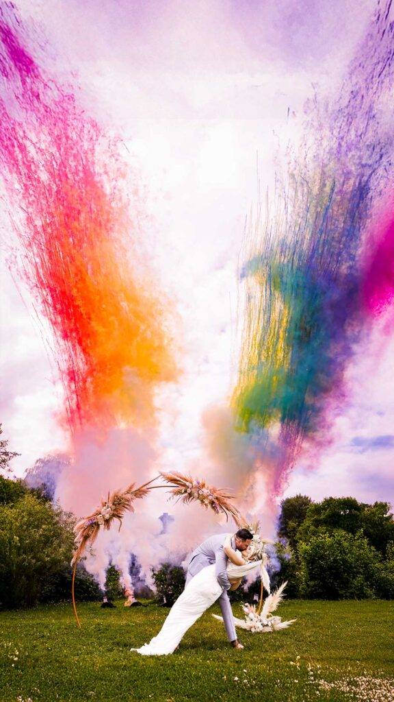 Ein Brautpaar küsst sich unter einem Traubogen auf Gut Groß Schwansee, während hinter ihnen riesige, bunte Rauchbomben explodieren und den Himmel in Regenbogenfarben färben. Ein spektakuläres Hochzeitsfoto.