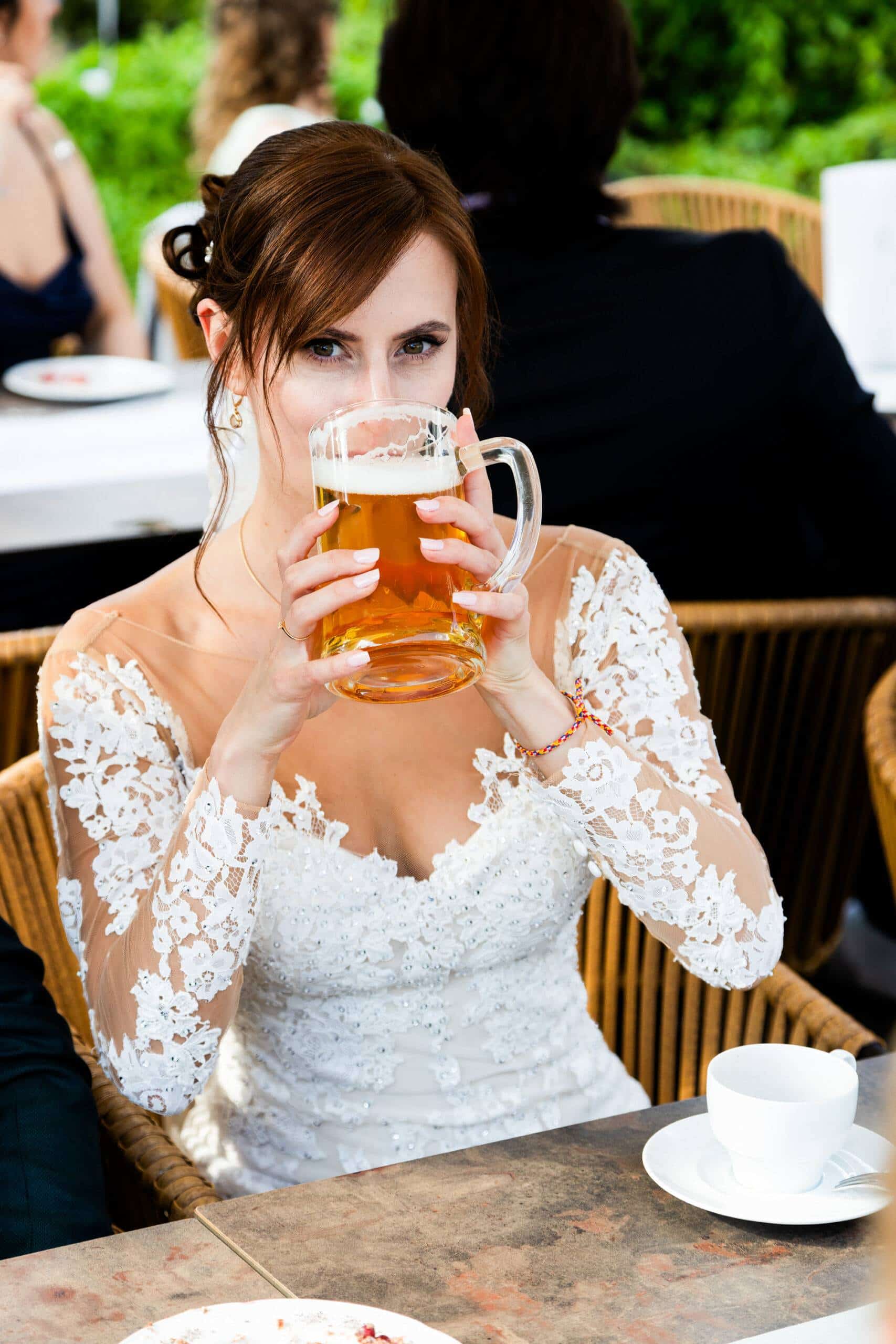 Eine Braut im Spitzenkleid sitzt entspannt an einem Tisch und trinkt mit einem Augenzwinkern Bier aus einem großen Glaskrug. Ein lustiger und authentischer Moment von der Hochzeitsfeier.