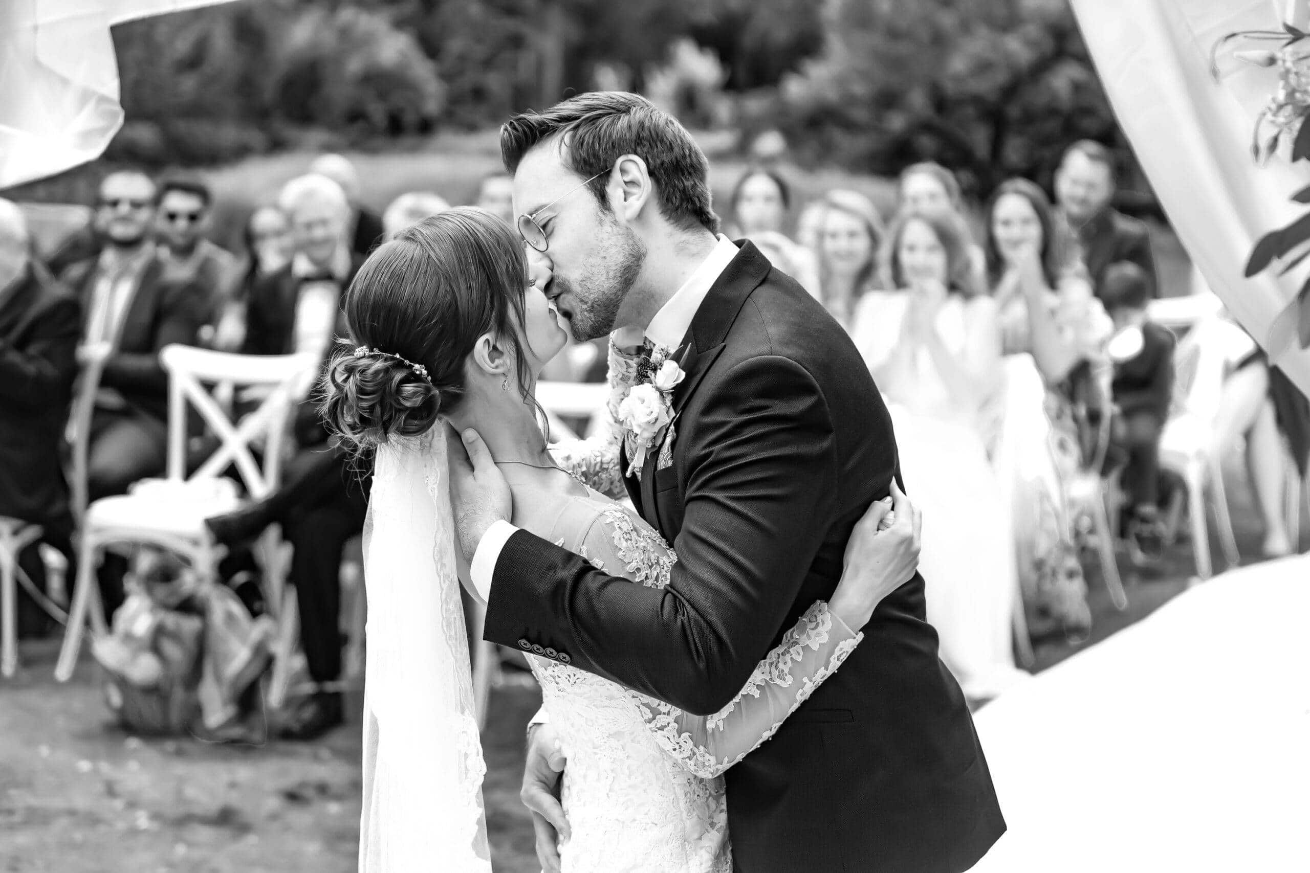 Das Brautpaar teilt seinen ersten Kuss als Ehepaar während einer Trauung im Freien. Im Hintergrund klatschen die Hochzeitsgäste. Schwarz-Weiß-Foto.