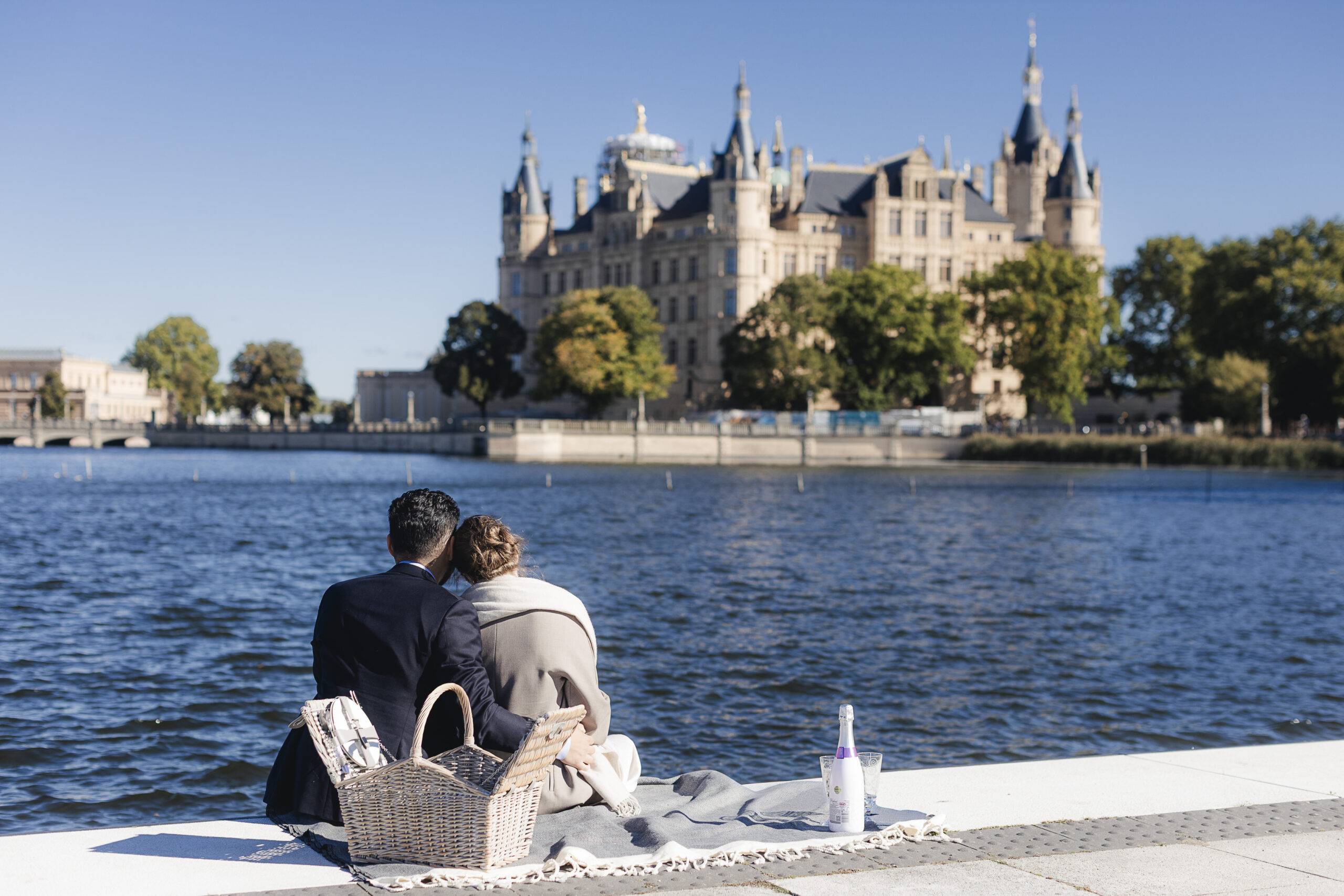 verlobung-schwerin-paar-blickt-auf-schloss Ein frisch verlobtes Paar sitzt von hinten auf einer Decke am Ufer in Schwerin und blickt gemeinsam über den See auf das Schweriner Schloss.
